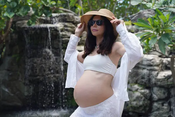 Hamile bir kadın zarif bir şekilde güzel bir şelale kayasının önünde duruyor..