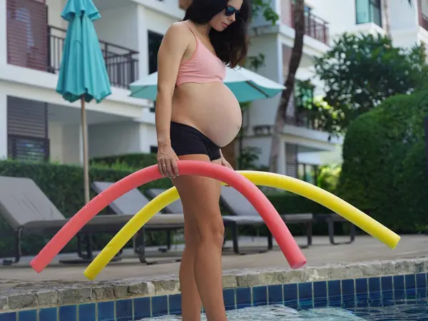 Bilardo oynamak üzere olan hamile genç kadın köpüklü makarnayla havuzda egzersiz yapacak..