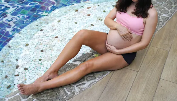 Pembe bikinili hamile bir kadın havuzun kenarında oturuyor..