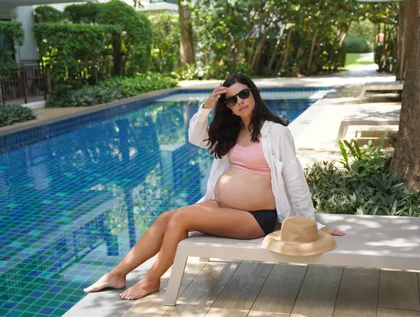 Pembe bikinili hamile bir kadın havuzun kenarında oturuyor..