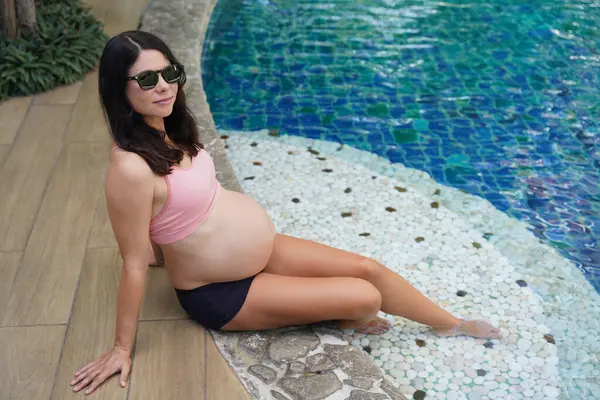 Pembe bikinili hamile bir kadın havuzun kenarında oturuyor..