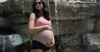 Hamile bir kadın zarif bir şekilde güzel bir şelale kayasının önünde duruyor..
