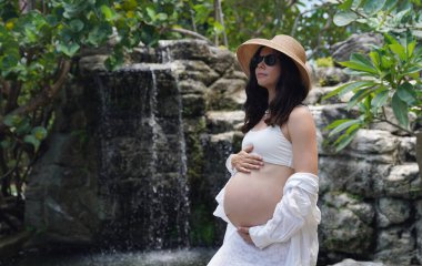Hamile bir kadın zarif bir şekilde güzel bir şelale kayasının önünde duruyor..