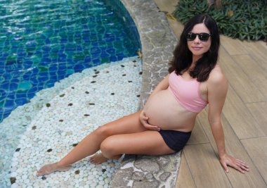 Pembe bikinili hamile bir kadın havuzun kenarında oturuyor..