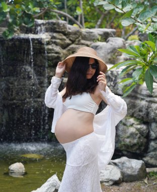 Hamile bir kadın zarif bir şekilde güzel bir şelale kayasının önünde duruyor..