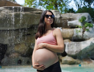 Hamile bir kadın zarif bir şekilde güzel bir şelale kayasının önünde duruyor..