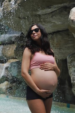 Hamile bir kadın zarif bir şekilde güzel bir şelale kayasının önünde duruyor..