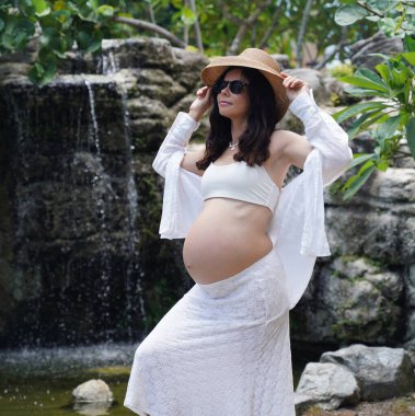 Hamile bir kadın zarif bir şekilde güzel bir şelale kayasının önünde duruyor..