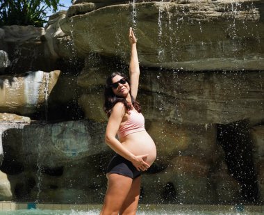 Hamile bir kadın zarif bir şekilde güzel bir şelale kayasının önünde duruyor..