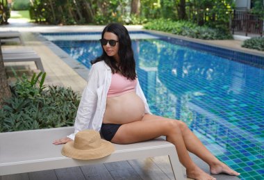 Pembe bikinili hamile bir kadın havuzun kenarında oturuyor..