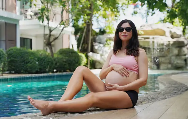 Pembe bikinili hamile bir kadın havuzun kenarında oturuyor..