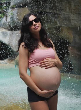 Hamile bir kadın zarif bir şekilde güzel bir şelale kayasının önünde duruyor..