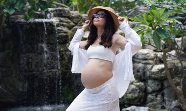 Hamile bir kadın zarif bir şekilde güzel bir şelale kayasının önünde duruyor..