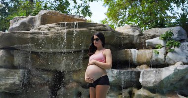 Hamile bir kadın zarif bir şekilde güzel bir şelale kayasının önünde duruyor..