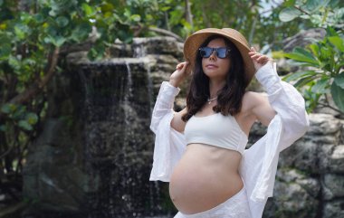 Hamile bir kadın zarif bir şekilde güzel bir şelale kayasının önünde duruyor..