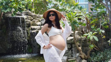 Hamile bir kadın zarif bir şekilde güzel bir şelale kayasının önünde duruyor..