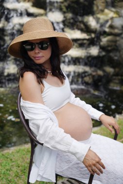 Hamile bir kadın zarif bir şekilde güzel bir şelale kayasının önünde oturuyor..