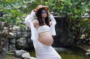 Hamile bir kadın zarif bir şekilde güzel bir şelale kayasının önünde duruyor..