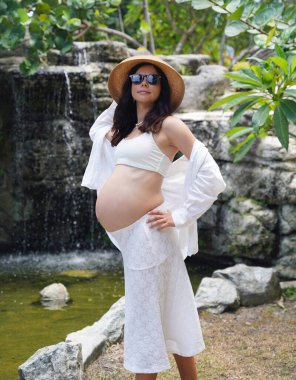 Hamile bir kadın zarif bir şekilde güzel bir şelale kayasının önünde duruyor..