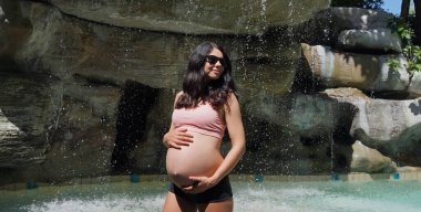 Hamile bir kadın zarif bir şekilde güzel bir şelale kayasının önünde duruyor..