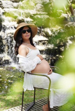 Hamile bir kadın zarif bir şekilde güzel bir şelale kayasının önünde oturuyor..