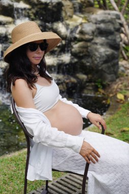 Hamile bir kadın zarif bir şekilde güzel bir şelale kayasının önünde oturuyor..