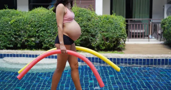 Bilardo oynamak üzere olan hamile genç kadın köpüklü makarnayla havuzda egzersiz yapacak..