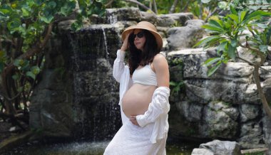Hamile bir kadın zarif bir şekilde güzel bir şelale kayasının önünde duruyor..