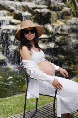 Hamile bir kadın zarif bir şekilde güzel bir şelale kayasının önünde oturuyor..