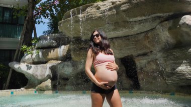 Hamile bir kadın zarif bir şekilde güzel bir şelale kayasının önünde duruyor..