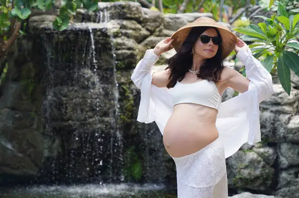 Hamile bir kadın zarif bir şekilde güzel bir şelale kayasının önünde duruyor..