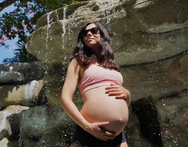 Hamile bir kadın zarif bir şekilde güzel bir şelale kayasının önünde duruyor..