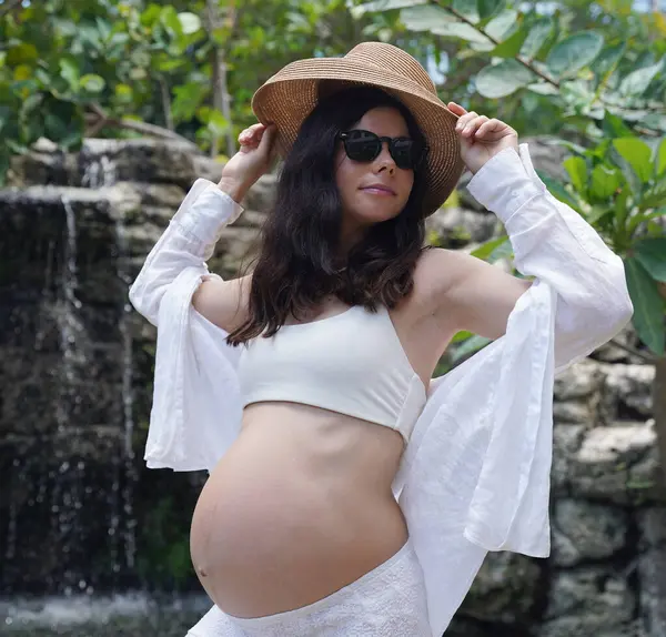 Hamile bir kadın zarif bir şekilde güzel bir şelale kayasının önünde duruyor..