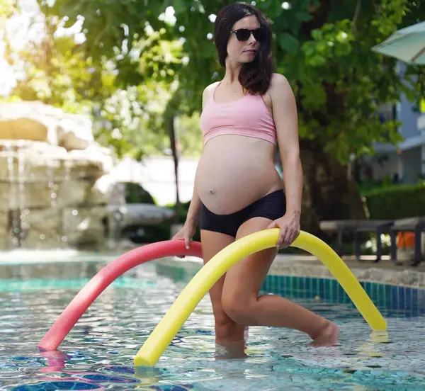 Bilardo oynamak üzere olan hamile genç kadın köpüklü makarnayla havuzda egzersiz yapacak..
