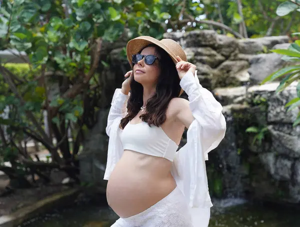 Hamile bir kadın zarif bir şekilde güzel bir şelale kayasının önünde duruyor..