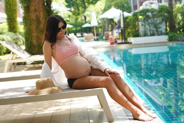 Pembe bikinili hamile bir kadın havuzun kenarında oturuyor..