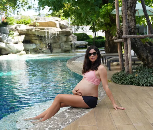Pembe bikinili hamile bir kadın havuzun kenarında oturuyor..