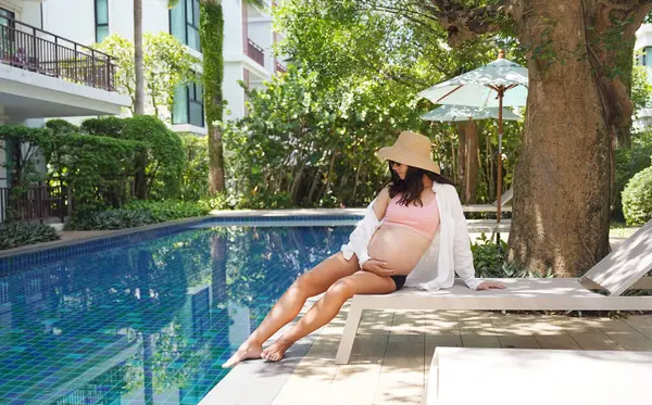 Pembe bikinili hamile bir kadın havuzun kenarında oturuyor..