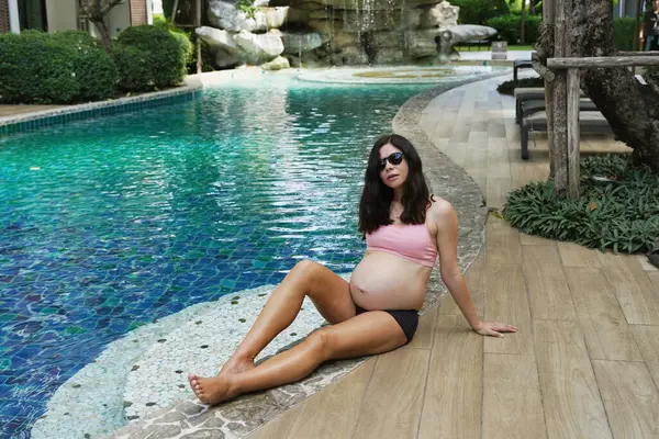 Pembe bikinili hamile bir kadın havuzun kenarında oturuyor..