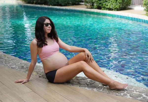 Pembe bikinili hamile bir kadın havuzun kenarında oturuyor..
