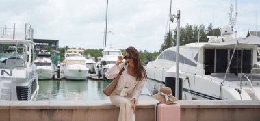 Pembe bavullu bir kadın, yat limanının önündeki beton bir çitin üzerinde oturuyor..