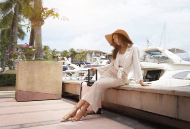 Pembe bavullu bir kadın, yat limanının önündeki beton bir çitin üzerinde oturuyor..