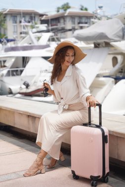 Pembe bavullu bir kadın, yat limanının önündeki beton bir çitin üzerinde oturuyor..