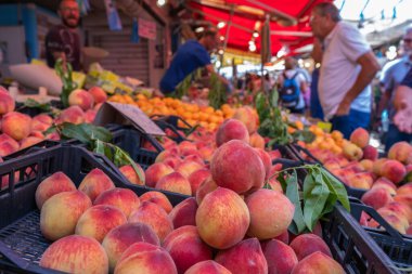 Şeftali tezgahta, arka planda gıda pazarında meyve satılıyor.