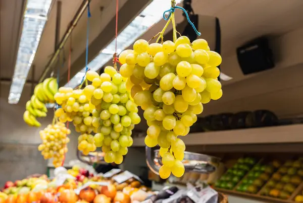 Yerel gıda pazarında bir meyve tezgahının üzerinde asılı duran beyaz üzüm kümeleri.