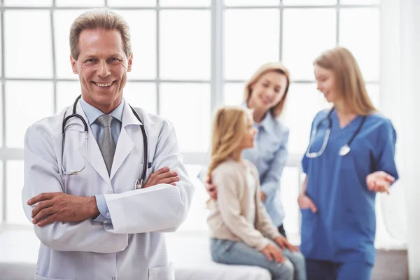 穿白色上衣的英俊中间岁儿科医生是站着双臂交叉和微笑 年轻的医生正在给病人的背景 — 图库照片