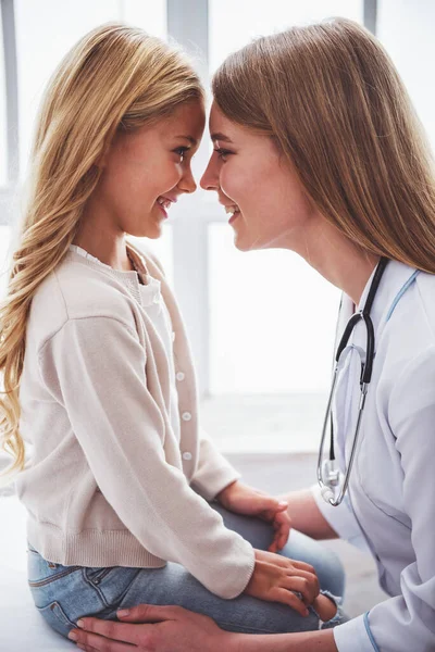 美丽年轻的儿科医生和可爱的小女孩触摸鼻子和微笑 坐在医生办公室 — 图库照片