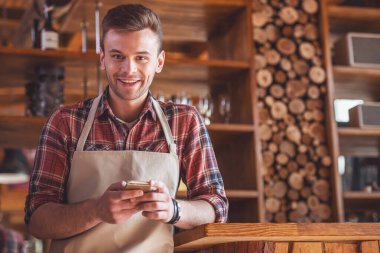 Yakışıklı genç barista önlük kameraya bakarak ve bara yaslanmış iken gülümseyen bir smartphone kullanarak sayaç Café