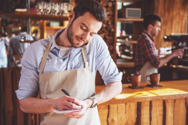 Yakışıklı genç garson cep telefonda konuşuyor ve notlar, arka plan barista yapma kahve yapıyor