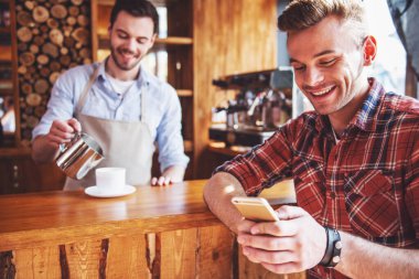Yakışıklı genç bir smartphone kullanarak ve gülümseyen, barda duran kahve, süt ekleyerek arka plan barista olarak sayaç